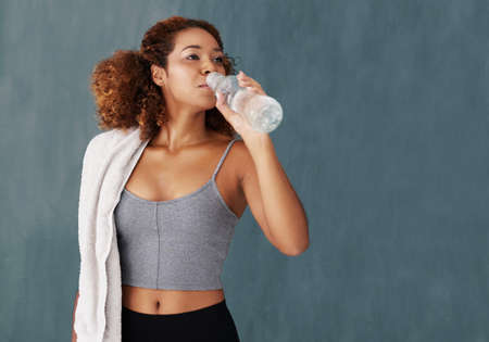 Water is an important part of any workout. Studio shot of a young woman drinking some water after yoga class against a grey background.の写真素材