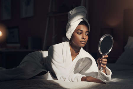 Spoil yourself with an at home spa day. Shot of a young woman looking at herself in the mirror while going through her beauty routine at home.の写真素材