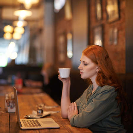 Relaxing with a fresh cup of coffee. Cropped shot of a young woman sitting with her laptop at a coffee shop.の写真素材