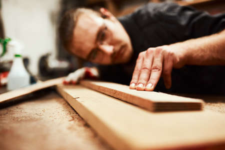 Thats the straightest its going to get. Cropped shot of a focused young male carpenter blowing dust off of a piece of wood after sanding it inside of his workshop during the night.の写真素材