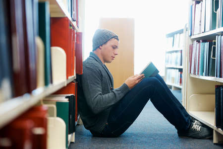 Getting his study material for the year. A young man sitting on the floor and reading in a library.