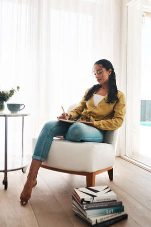 Note taking is how I remember all the important things. Full length shot of an attractive young businesswoman sitting alone in her living room and writing notes.の写真素材
