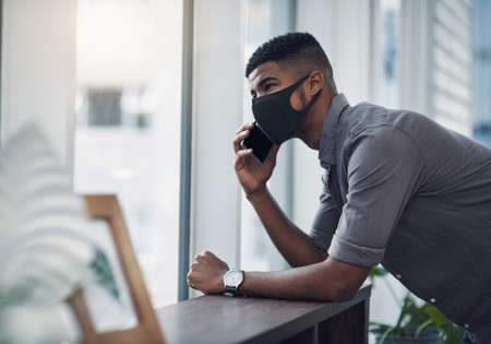 Business goes on, as long as we keep safe. Shot of a young businessman wearing a face mask while talking on a cellphone in an office.の写真素材