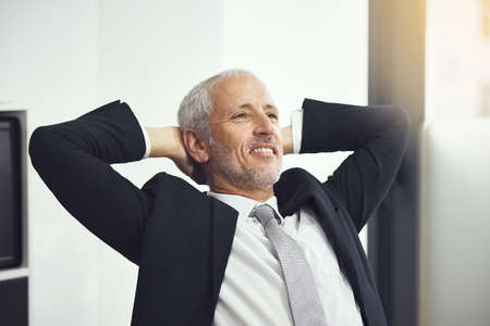 Taking care of business. Shot of a confident mature businessman leaning back in hais chair with his hands behind his head in an office.の写真素材