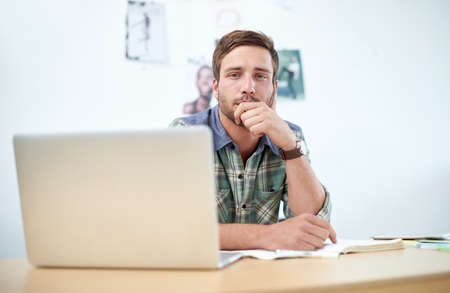 Portrait of a casually-dressed young man using a digital tablet at his desk. The commercial designs displayed in this image represent a simulation of a real product and have been changed or altered enough by our team of retouching and design specialists so that they are free of any copyright infringements.の写真素材