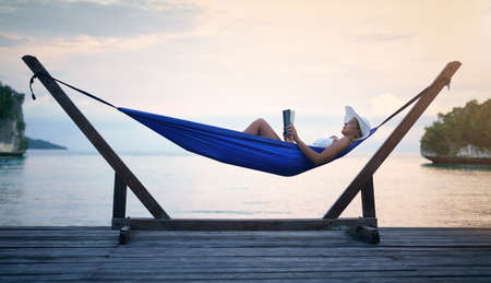 Shh, Im on vacation mode. Full length shot of an attractive young woman sitting alone in a hammock and reading a book during a vacation.の写真素材