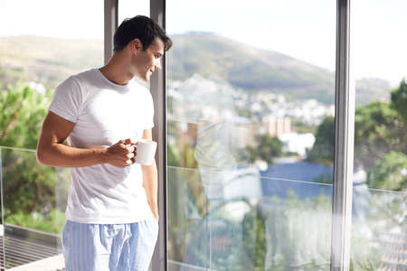 Waking up with fresh coffee and a great view. Cropped shot of a handsome young man drinking coffee while standing at his bedroom window.の写真素材