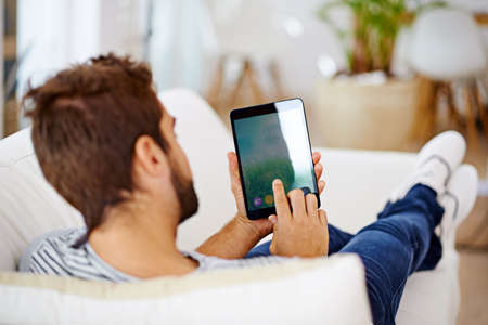 Enjoying some down-time with his latest tech. Rearview of a man using his tablet on his couch.