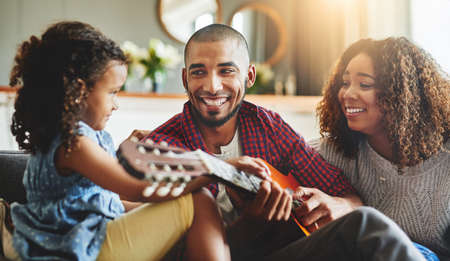 Tuned into family time. Shot of an adorable little girl and her parents playing a guitar together on the sofa at home.の写真素材