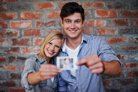 Creating memories together. A young couple standing in front of a brick wall holding a photo to the camera.の写真素材