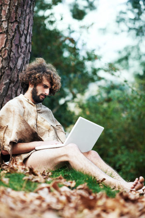 What scorcery is this. A confused looking caveman sitting by a tree with a laptop.