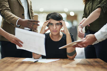 Does anyone have a headache tablet. Cropped portrait of a young businesswoman feeling overwhelmed by her colleagues in the office.の写真素材