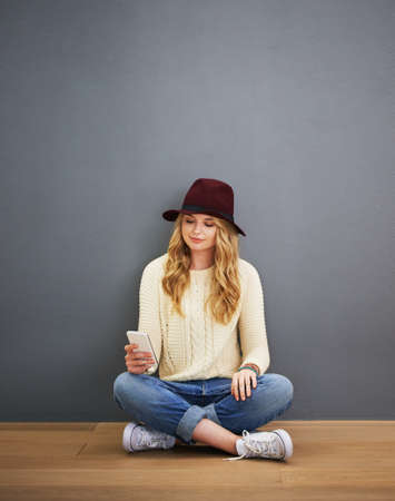 Reading a text. Shot of a young woman using her cellphone while sitting against a gray background.