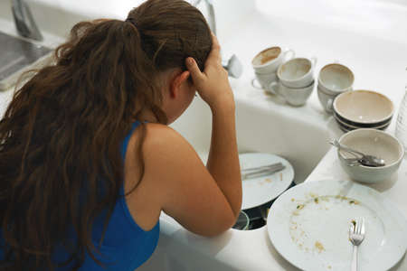 Shes had a long day. Shot of a frustrated looking woman standing by a pile of dirty dishes.の写真素材