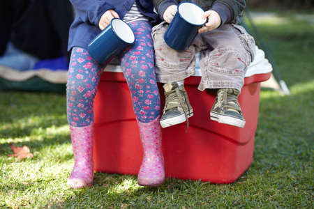 Tiny campers just relaxing. Cropped view of two small children holding tin mugs while sitting on a...