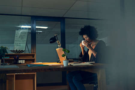 Shes got a strong cuppa to keep her going. Cropped shot of a young businesswoman working late on a laptop in an office.の写真素材