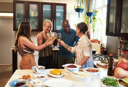 We are going to have a brilliant time. Cropped shot of a group of cheerful young friends having a celebratory toast with drinks while standing in the kitchen preparing food at home.の写真素材