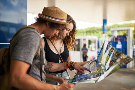 Lets take the scenic route. Shot of a young backpacking couple getting directions from a map at a gas station.の写真素材