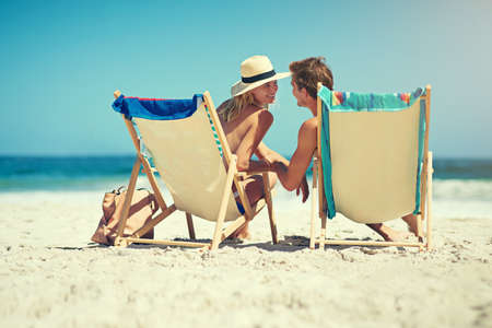 It just wouldnt be the same without you. Rearview shot of a young couple sitting on loungers at the beach.の写真素材