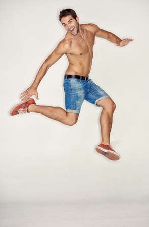 Filled with exuberance. Muscular young guy jumping in denim shorts while against a white background.の写真素材