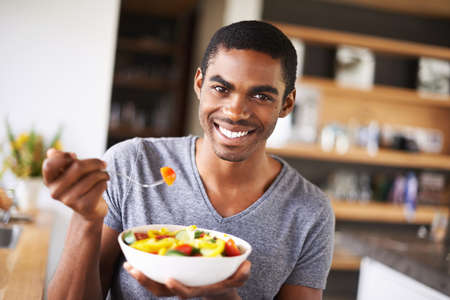 Have a bite of health. Indoor shot of a gorgeous young black man showing off his fruit salad.の写真素材