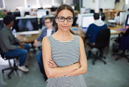 We bring ideas into reality. Portrait of a young woman standing in an office with designers in the background.の写真素材