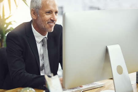This is very interesting indeed. Shot of a smiling mature businessman working on his computer in an...