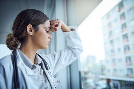 Burnout is rampant amongst medical professionals. Shot of a young female doctor looking stressed out while standing at a window in a hospital.の写真素材