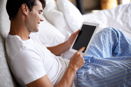 Watching movies in bed. Cropped shot of a handsome young man using his tablet in bed.の写真素材