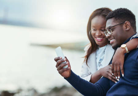 Check this out. Shot of an affectionate young couple using a cellphone together outdoors.の写真素材