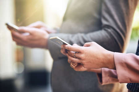 Download the app that connects you. Cropped shot of two people using their cellphones while standing outside.の写真素材
