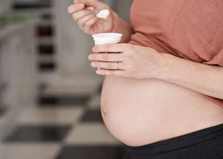Its a very healthy snack. Cropped shot of a young pregnant woman eating a yogurt in the kitchen.の写真素材