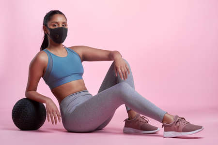 Im determined to reach my fitness goals. Full length portrait of an attractive and sporty young woman wearing a mask and posing with a medicine ball in studio against a pink background.の写真素材