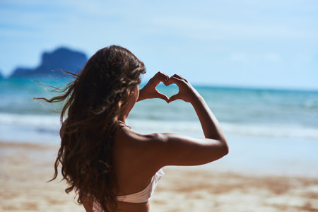 I just love the beach. Shot of a young woman shaping a heart while looking at the scenery at the beach.の写真素材