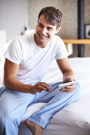 What should I do now. Cropped shot of a handsome young man using a tablet on the edge of his bed.の写真素材