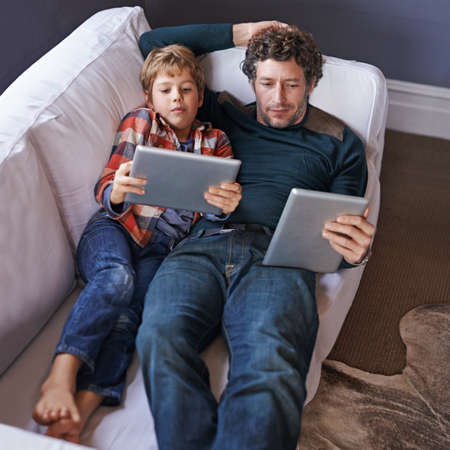 Like father, like son. Shot of a father son lying on the couch together while using digital tablets.の写真素材