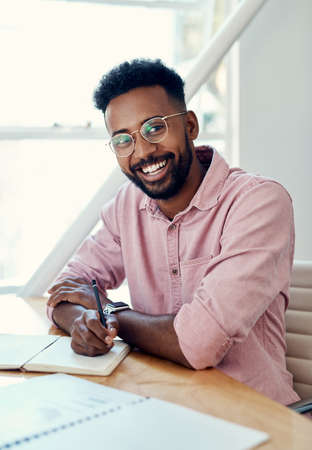 Note-taking is so undervalued these days. Cropped portrait of a handsome young businessman sitting and writing in a notebook while in the office alone.の写真素材