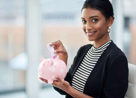 Even a little bit goes a long way in business. Portrait of an attractive young businesswoman putting money inside her piggy bank at work.の写真素材