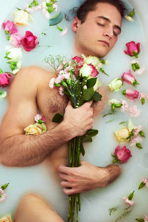 Its not a relaxing bath if the water is not milky. High angle shot of a handsome young man holding a bouquet while lying in a bathtub full of milky water and flower petals at home.の写真素材