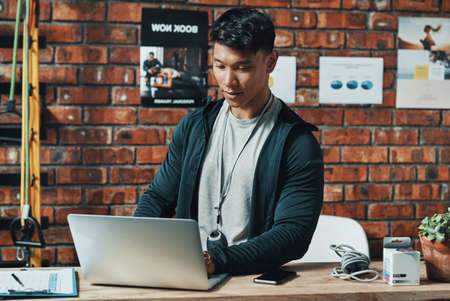 Hes receiving even more subscriptions online. Cropped shot of a handsome young male fitness instructor using a laptop while working in a gym.の写真素材