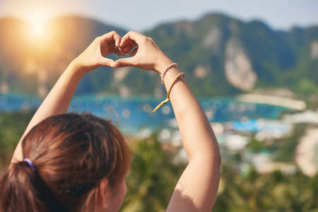 This island has stolen her heart. Rearview shot of a happy young tourist making a heart with her hands against a beautiful landscape.の写真素材