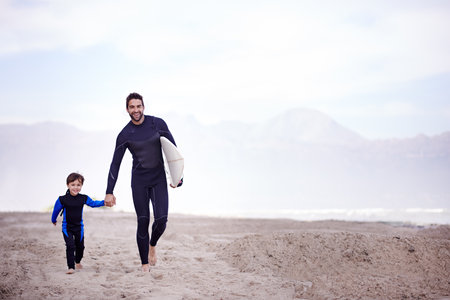 The waves are waiting, lets go. A father walking along the beach with his young son before a surfing...