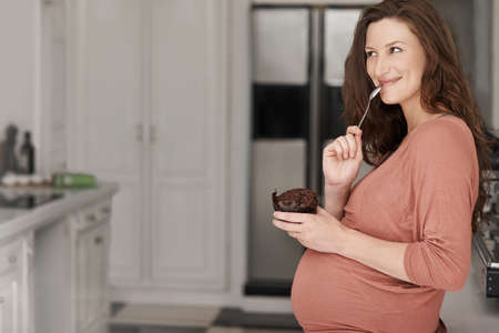 One cupcake couldnt hurt.... Cropped shot of a young pregnant woman eating a cupcake in the kitchen.