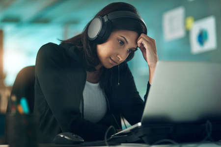 On the bench cos my confidence wont let me fly. Portrait of an exhausted young woman using a headset in a modern office.の写真素材