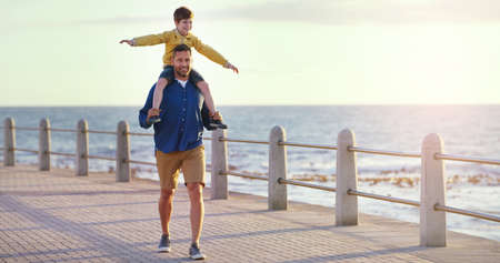 Flying high in the sky. Shot of a cheerful father carrying his son on his shoulders while taking a walk on walkway near the beach.の写真素材