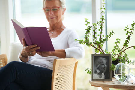 Relaxed and retired. A senior woman reading a great book in her lounge alongside an old photograph of herself.の写真素材