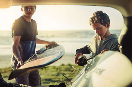 We surf, therefore we are. Shot of two young brothers unloading their surfboards from the back of a car by the beach.の写真素材