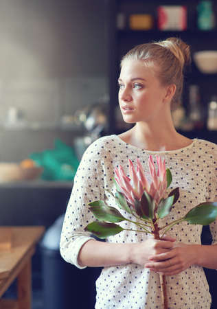 Floral inspiration. Shot of a beautiful young woman standing in her kitchen holding a protea flower.の写真素材