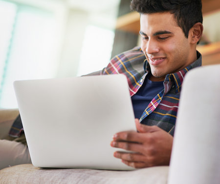 Spending a little time online. Shot of a handsome young man using his laptop while relaxing on the sofa at home.の写真素材