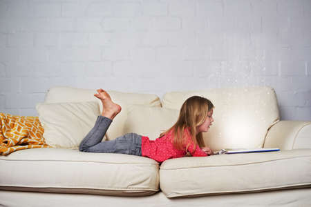 Theres so much magic in reading. Shot of a little girl reading a book while lying on a sofa.